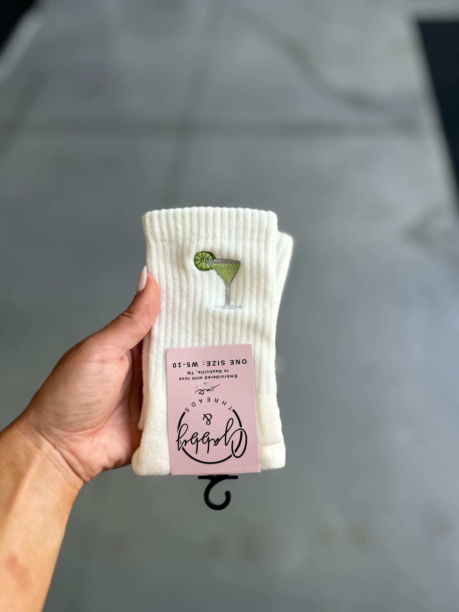Hand holding a pack of white socks with an embroidered margarita against a gray background