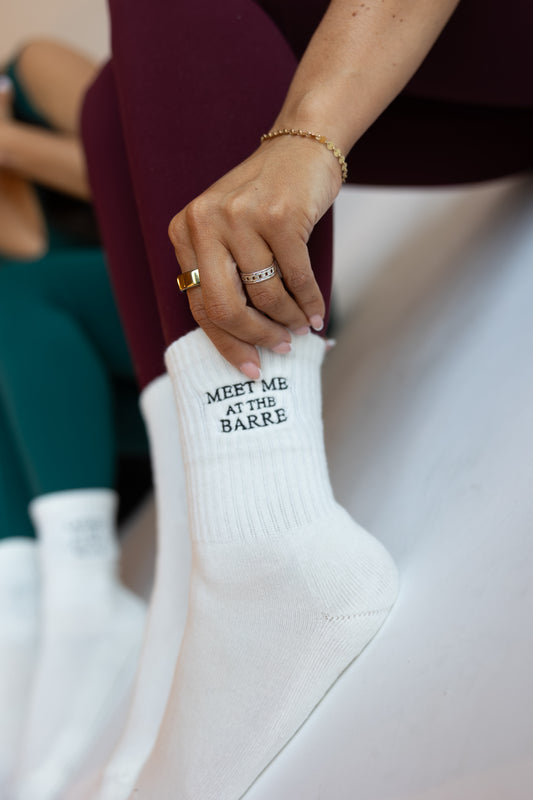 White sock with 'MEET ME AT THE BARRE' text worn by a person sitting on a white surface.