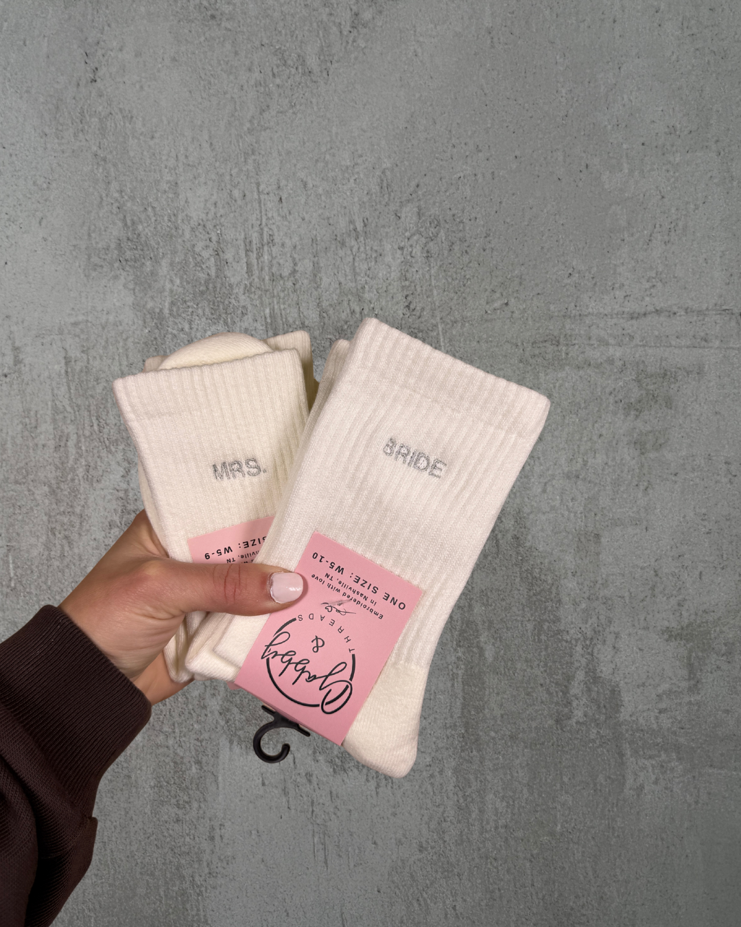 a hand holds up two pairs of socks, one with the word "bride" and the other with the word "Mrs." against a gray background