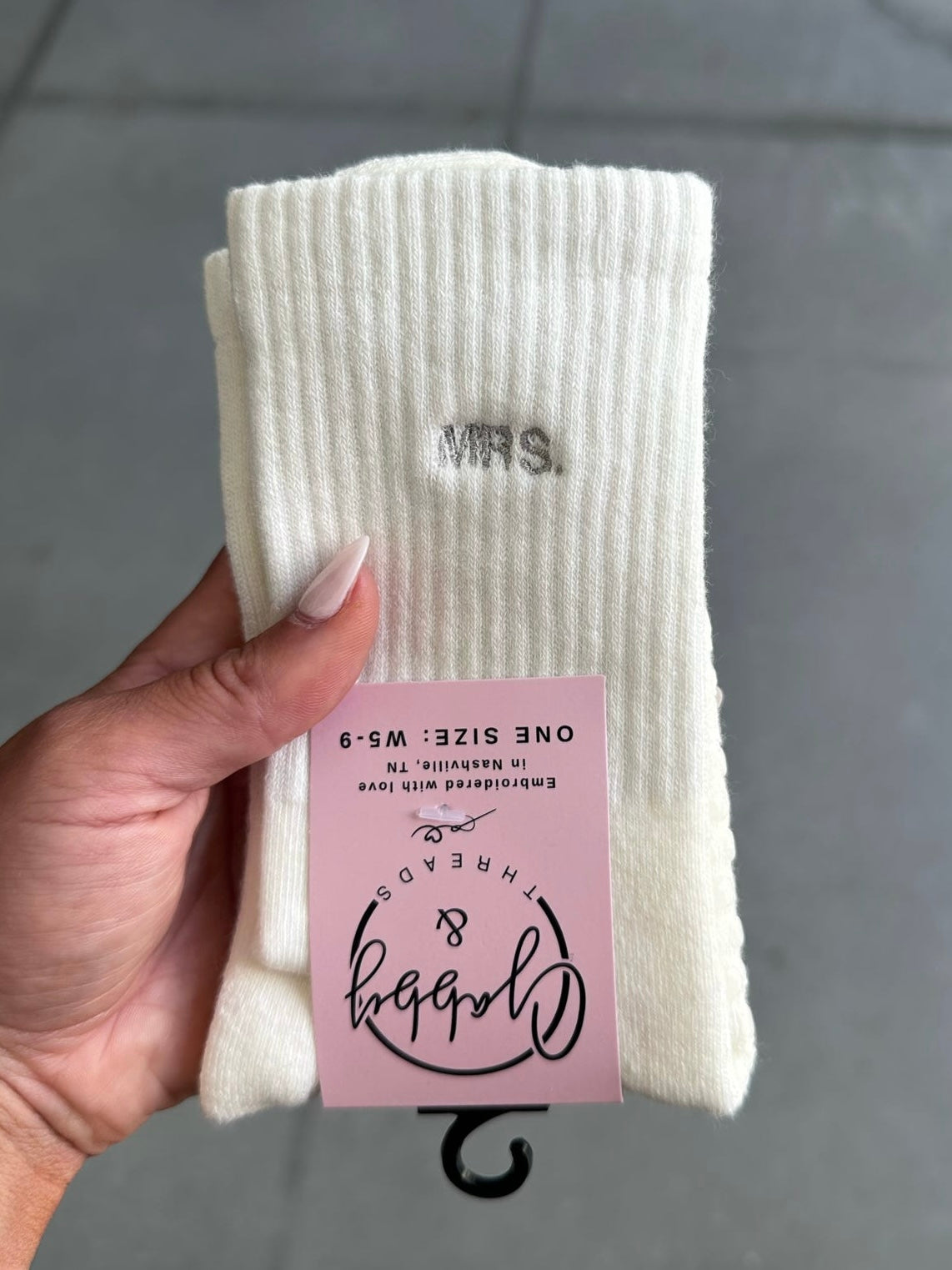 White socks with "MRS." embroidered with a pink label held by a hand against a gray background