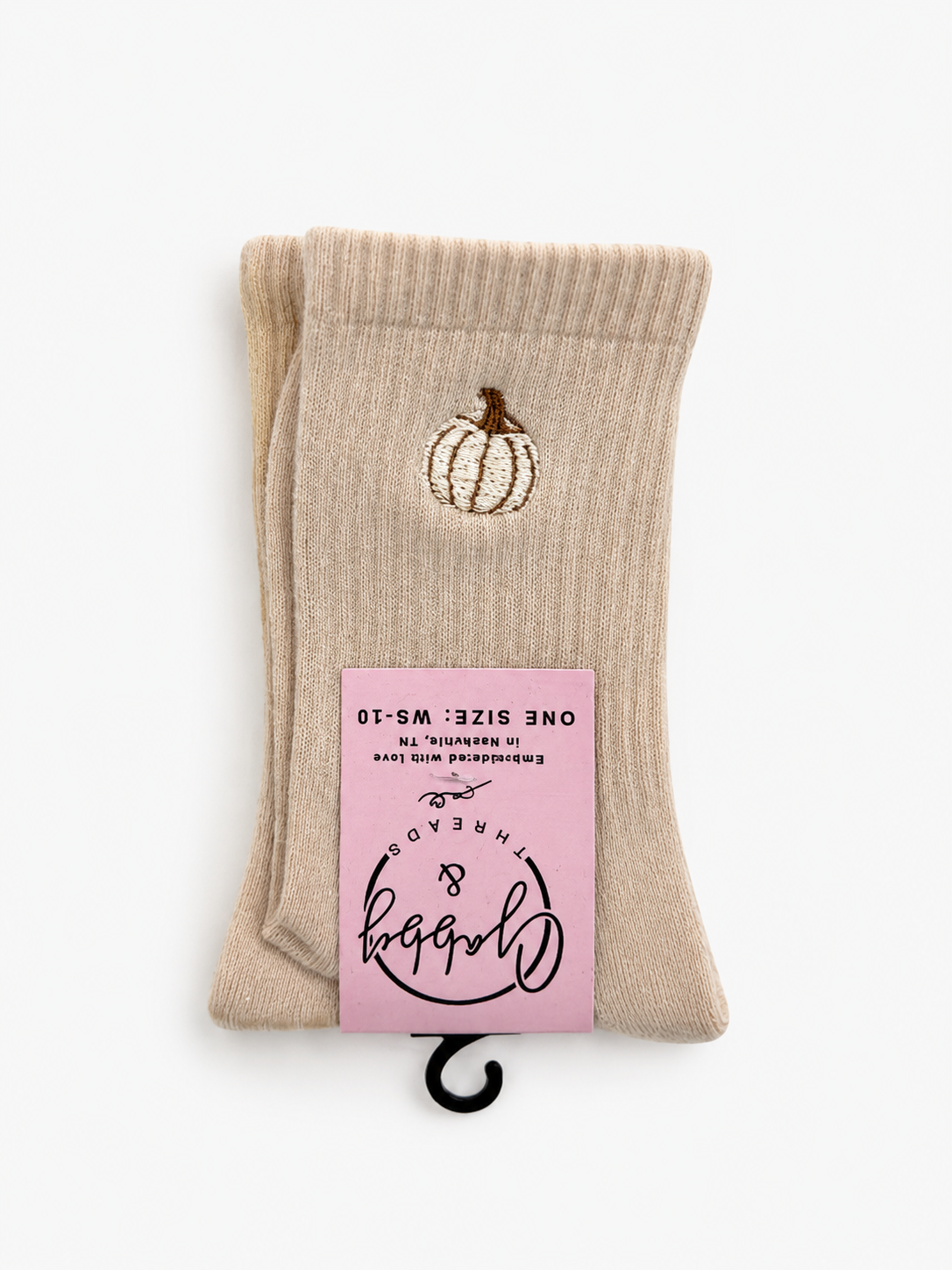 A pair of beige ribbed crew socks folded together with a pink paper tag attached. Each sock has a small embroidered pumpkin design near the top.