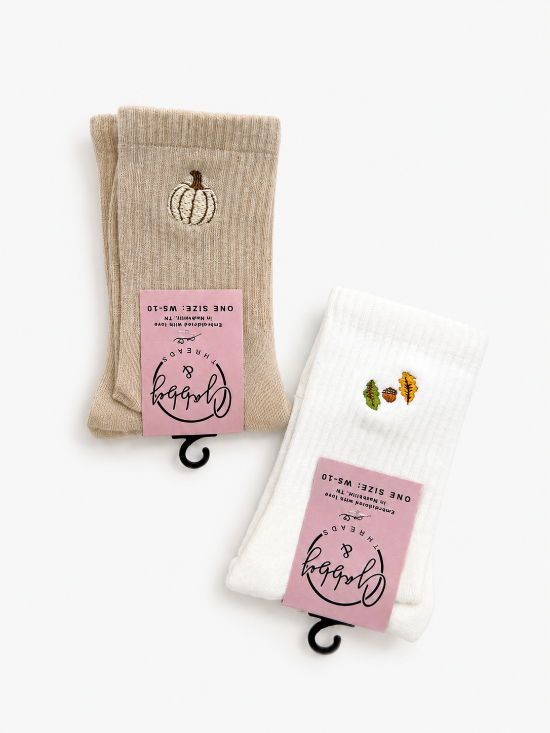 Two pairs of ribbed crew socks on black hangers against a white background. One pair is beige with a small embroidered pumpkin motif, and the other is white with a small embroidered multicolor leaf motif.