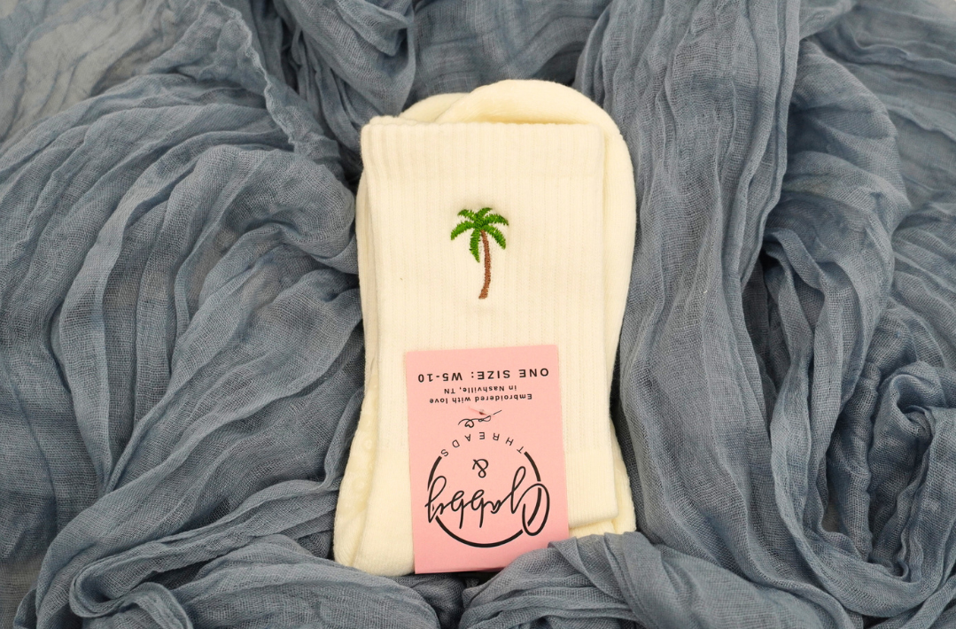 a pair of white grip socks with a palm tree embroidered on them lay against a blue background