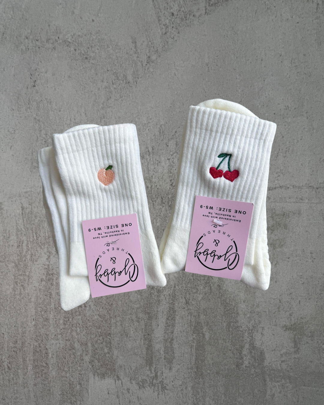 two pairs of white grip socks with embroidery on them. One has an embroidered peach and the other one has embroidered cherries on them 