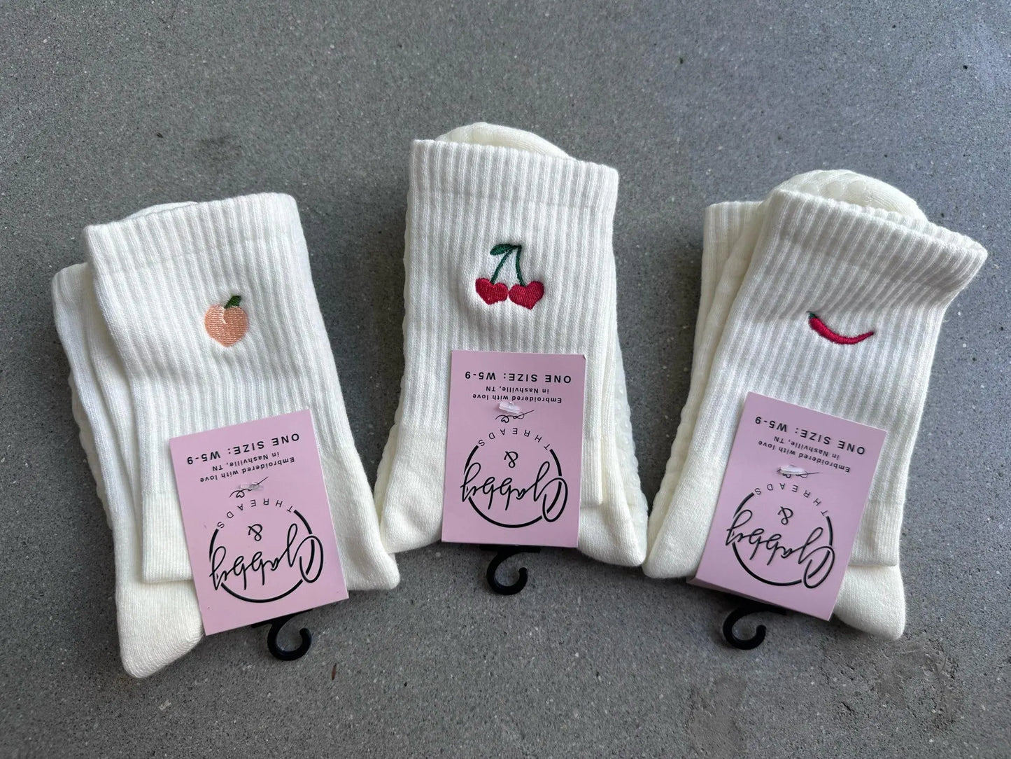 Three pairs of crew socks are laying on a cement floor. One pair has a peach embroidered on it, one has a red pepper embroidered on it and the other one has cherries embroidered on it.