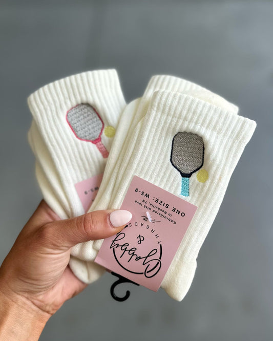 Hand holding two pairs of white socks with embroidered pickleball paddles against a gray background