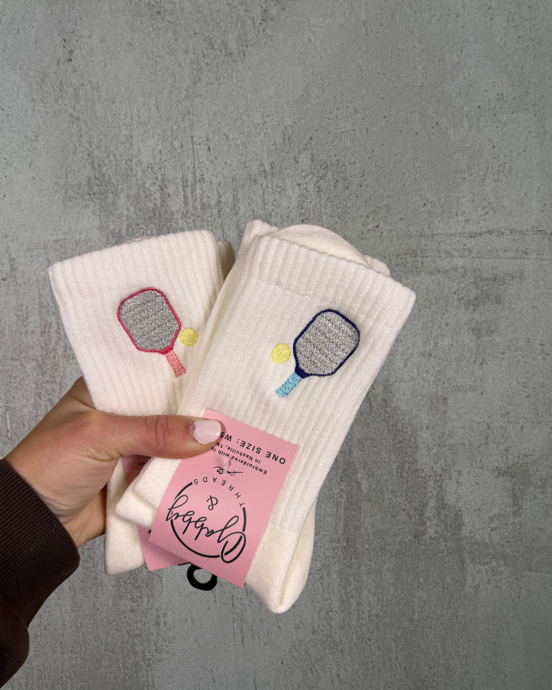 Hand holding two pairs of white socks with embroidered pickleball paddles against a gray background