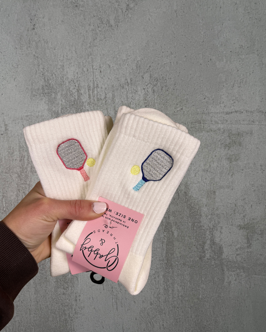 Hand holding two pairs of white socks with embroidered pickleball paddles against a gray background