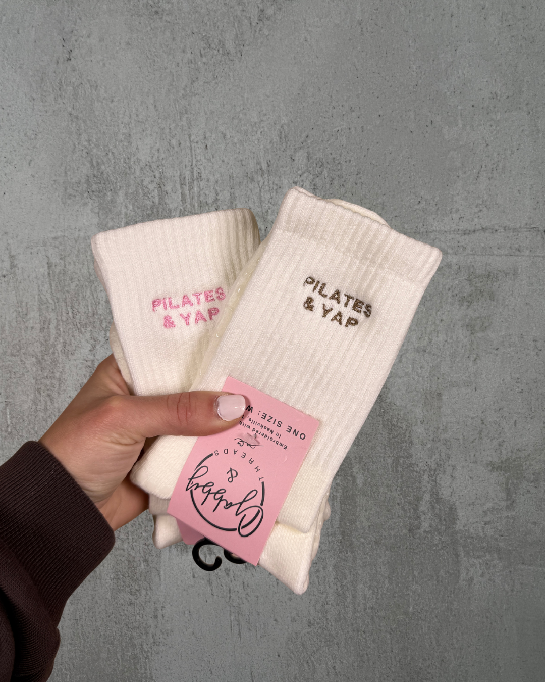 a hand holds up two pairs of socks. Both pairs of socks have the text "pilates & yap" embroidered on them against a gray background