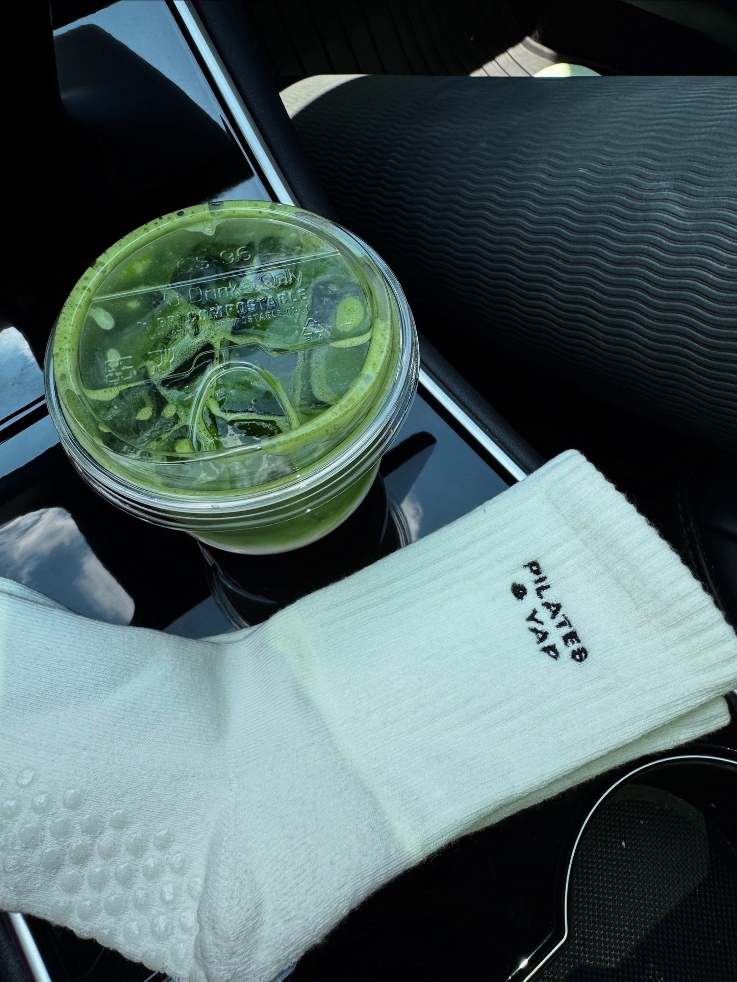 a pair of white grip socks is laying across a car console. The socks have the words "pilates and yap" embroidered on them. There is a green matcha tea in the cup holder