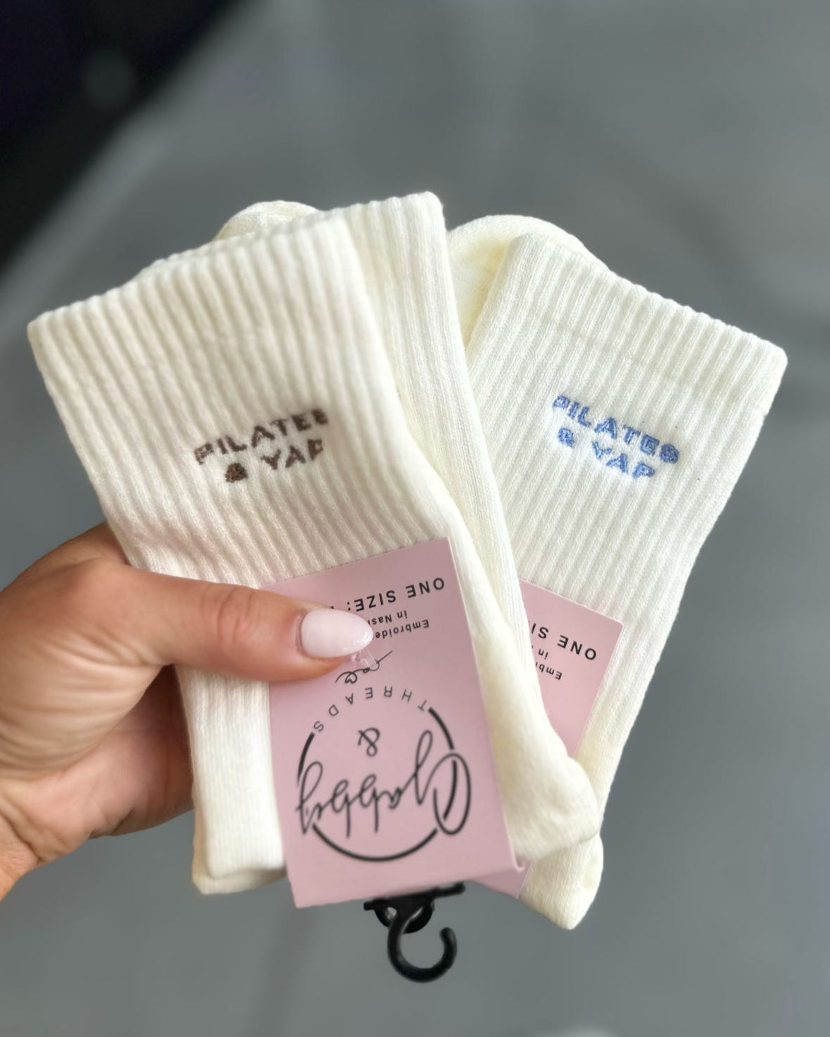White grip socks with "Pilates & Yap" held by a hand on a blurred background