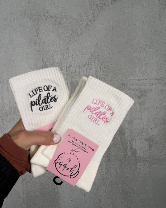 a hand holds up two pairs of socks, both pairs of socks have the words "life of a pilates girl" embroidered on them against a gray background
