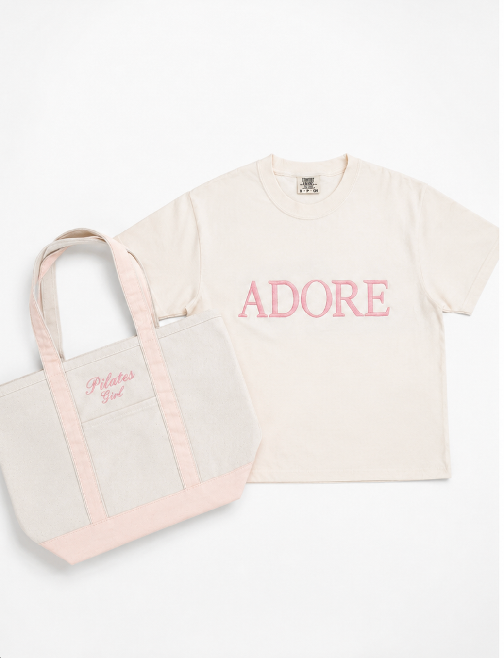 a pink canvas tote bag with the words "pilates girl" lays next to an embroidered cream colored shirt that says "adore"