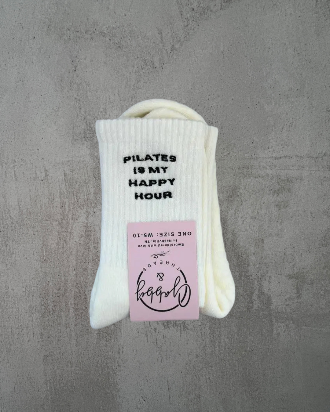 White grip socks with 'Pilates is my happy hour' text embroidered on against a gray background