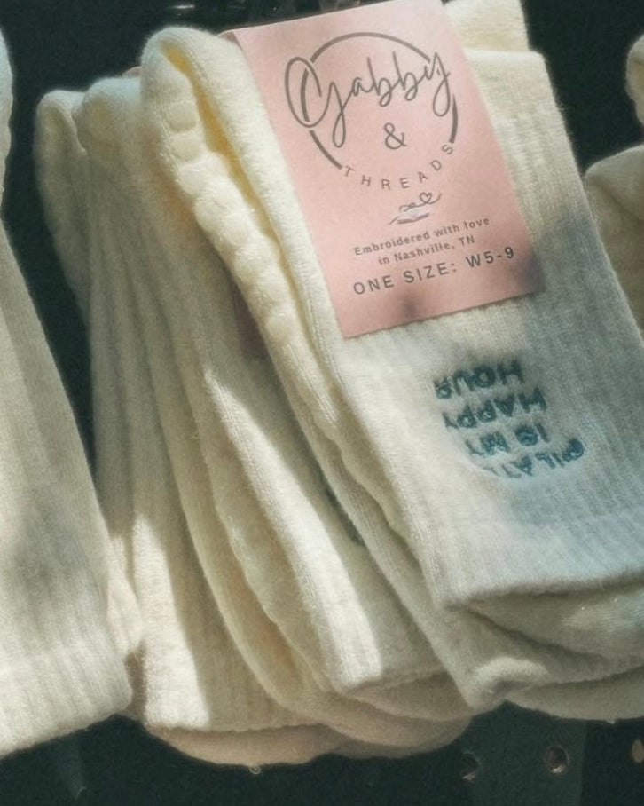Pair of grip socks with the text "pilates is my happy hour" are hanging on a peg board