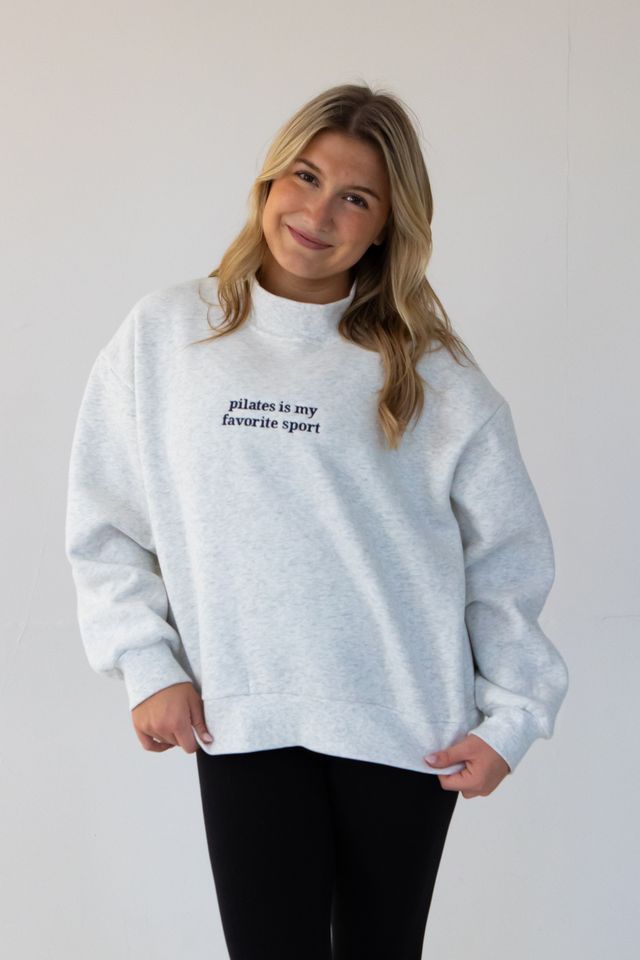 a model is wearing a heather gray mockneck that has "pilates is my favorite sport" embroidered on it