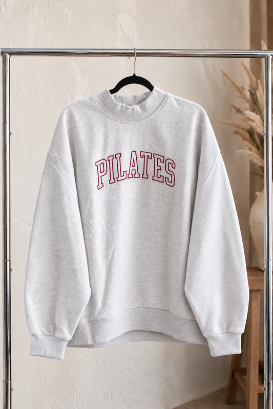 a heather gray mock neck sweatshirt is hanging on a clothing rack. The sweatshirt is embroidered with the word "PILATES" in varsity letters