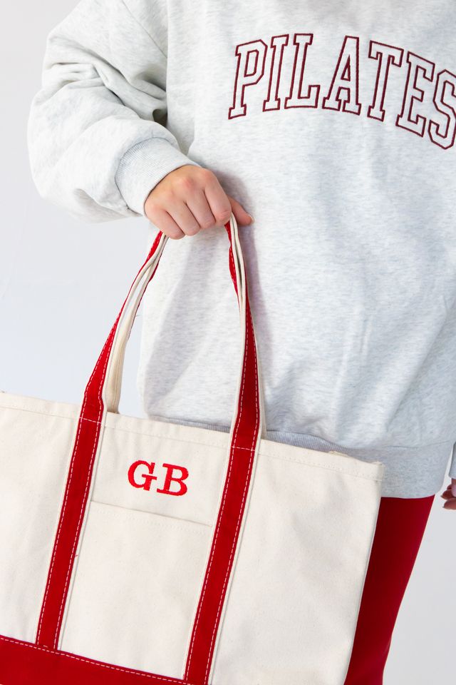 a woman is wearing a heather gray mock neck with the words "pilates" embroidered on it in varsity font while holding a red canvas tote bag