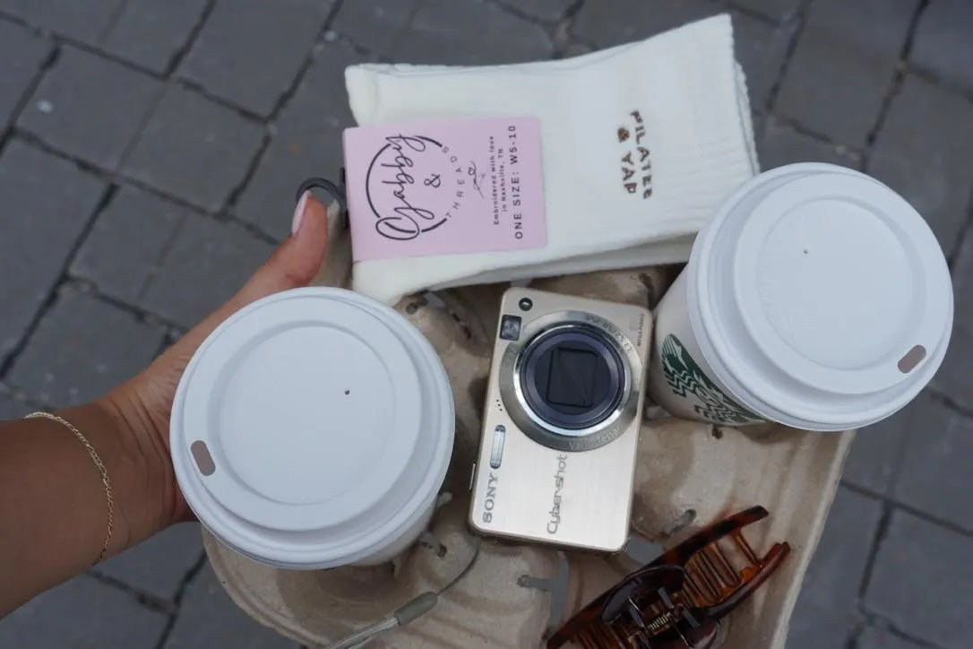 a coffee drink tray has two coffee cups, a hair clip, digital camera and a white pair of grip socks with the text "pilates and yap" embroidered on them