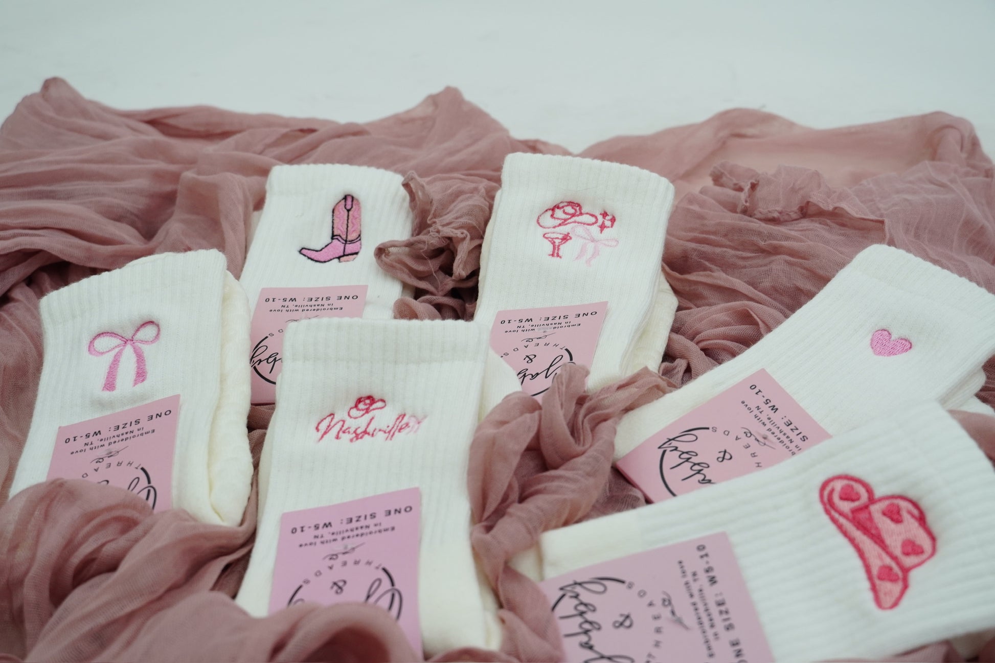 a collection of white grip socks with different pink embroidered icons laying on a pink tablecloth