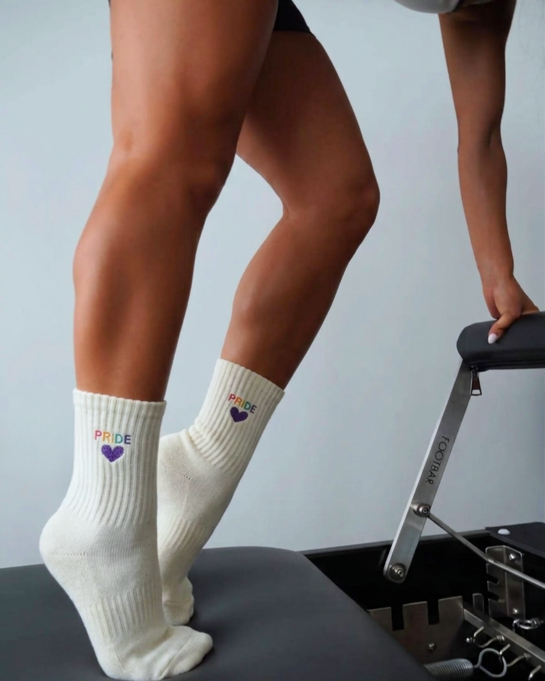 a person is standing on a pilates reformer wearing a pair of grip socks that say "PRIDE" in rainbow thread colors
