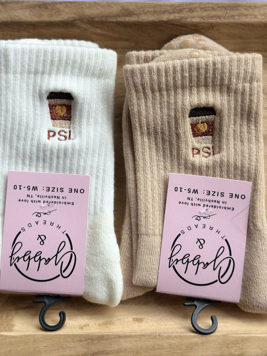 Two pairs of socks on a wooden tray with pink tags displaying a pumpkin spice latte with the letters PSL
