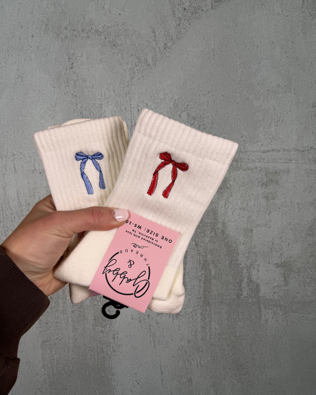 Two pairs of socks with embroidered bows held by a hand against a gray background