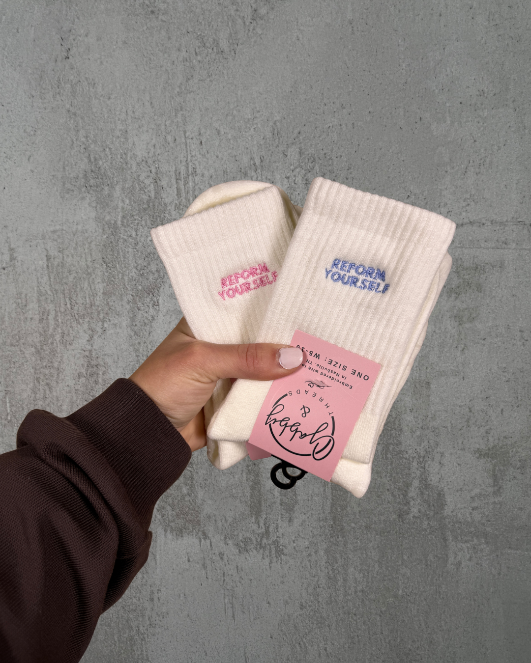 two pairs of white grip socks with the text "reform yourself" embroidered on them held against a grey wall