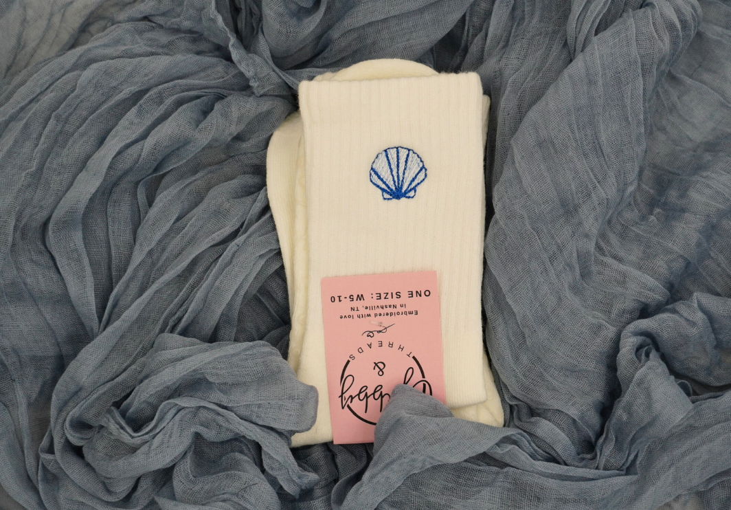 a pair of white grip socks with an embroidered seashell against a blue background