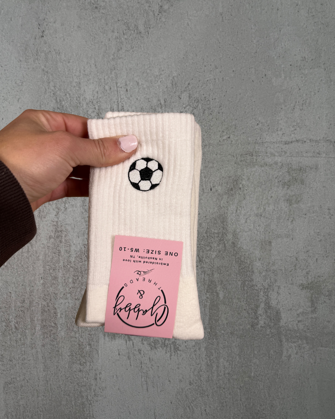a hand holds up a pair of white crew socks with an embroidered soccer ball on it against a grey wall.
