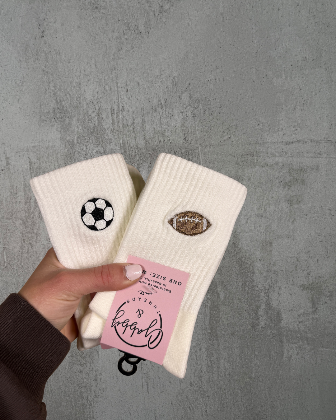 a hand holds up two pairs of white crew socks. One has an embroidered soccer ball on it and the other an embroidered football. They are held up against a grey background