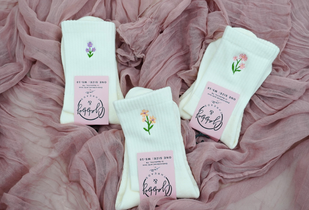 Three white socks with different colored flowers embroidered on them laying on a pink textured fabric background