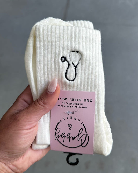 White crew socks with a black and gray stethoscope design held by a hand against a gray tiled floor background