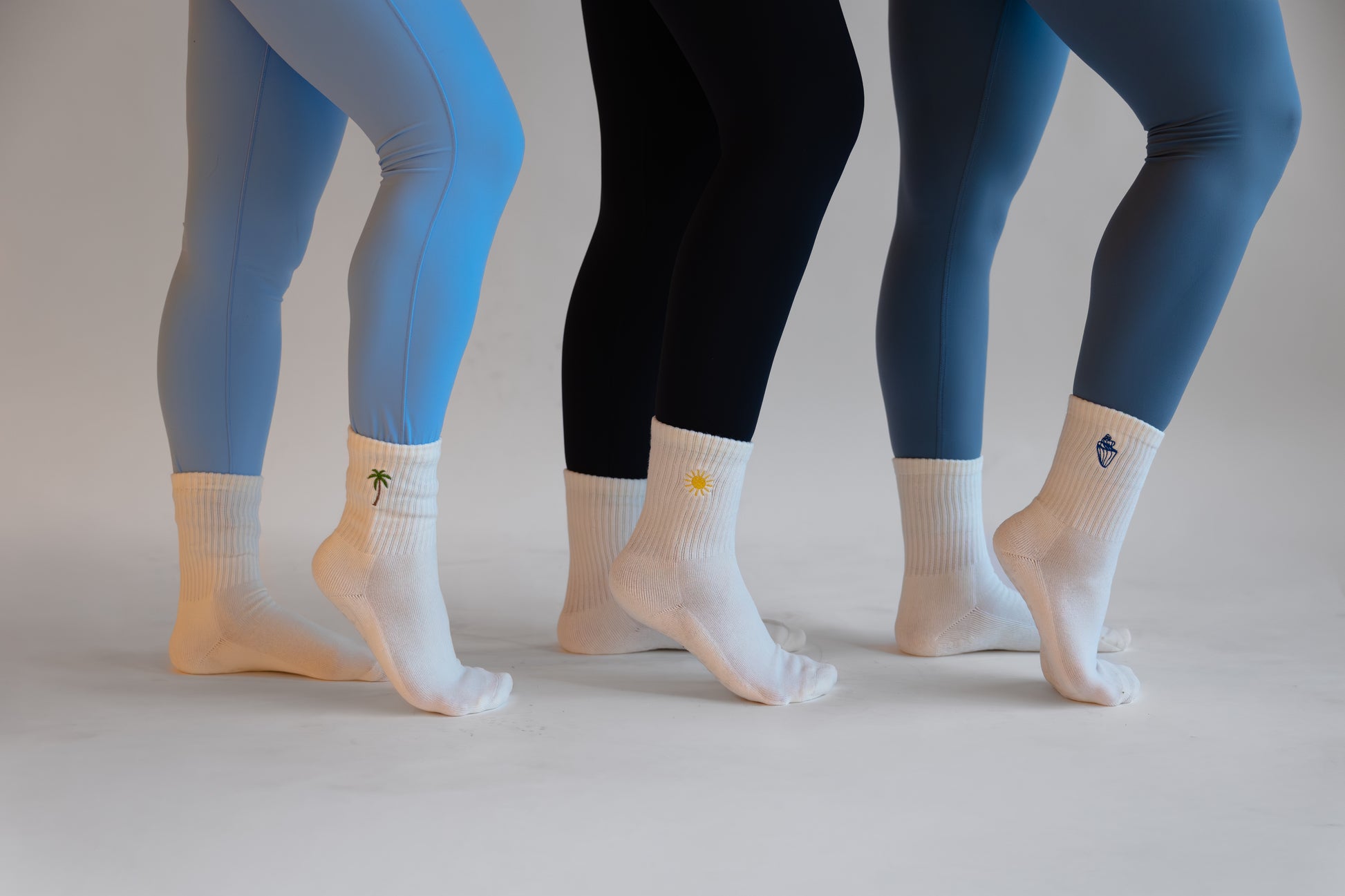 Three pairs of legs wearing different colored leggings and socks on a neutral background. The socks have different embroidered icons (palm tree, sun, and conch shell)