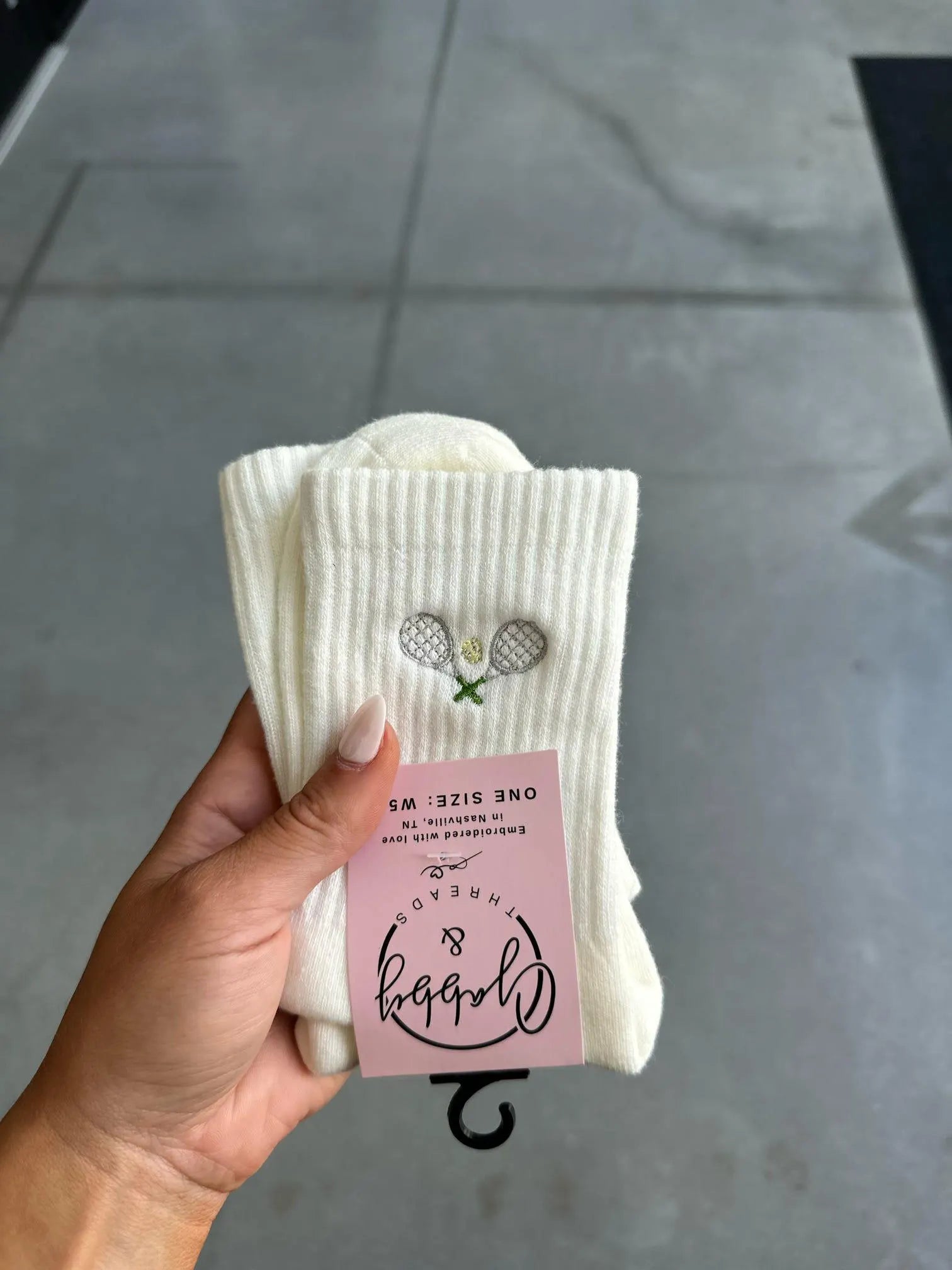 White crew socks with an embroidered tennis racket on them held up by a hand against a grey background