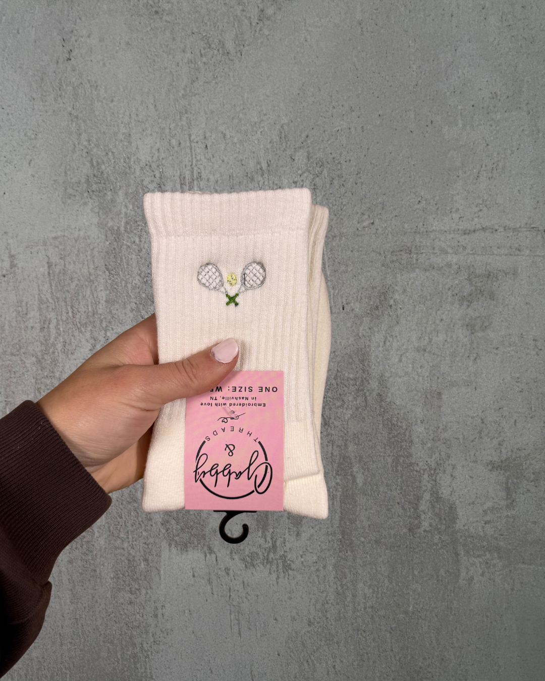 White crew socks with an embroidered tennis racket on them held up by a hand against a grey background