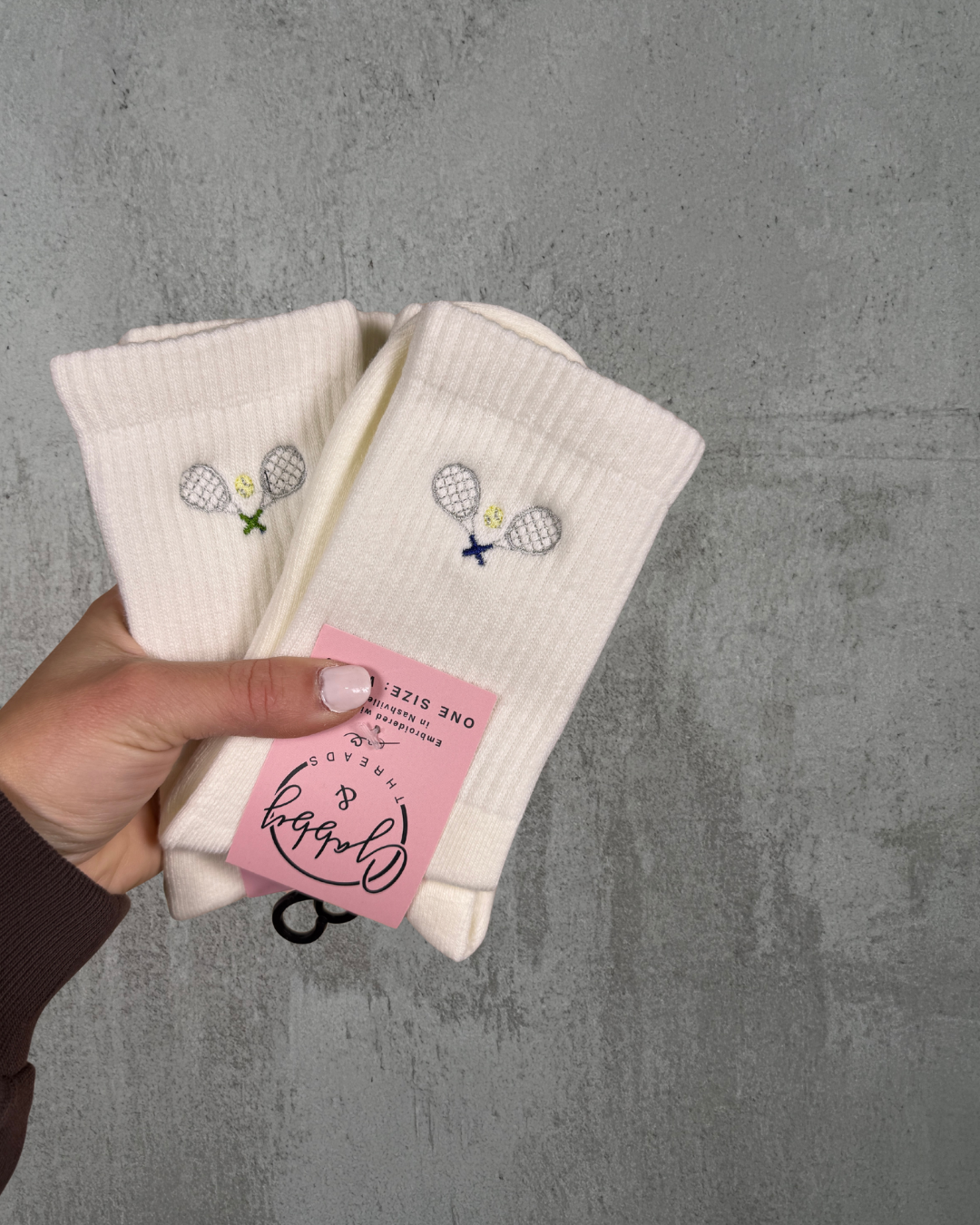 two pairs  of white crew socks with an embroidered tennis racket on them held up by a hand against a grey background