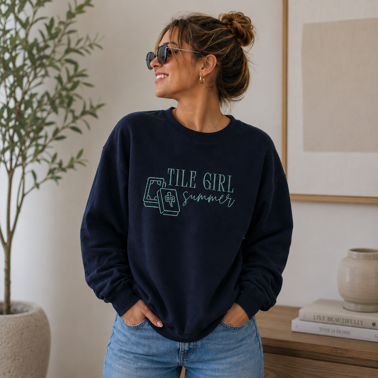 A woman wearing a navy blue crewneck sweatshirt with 'TILE GIRL summer' and  mahjong tiles embroidered on the front, paired with light wash blue jeans in a bright indoor setting.