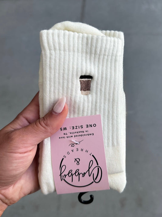White socks with to-go coffee cups embroidered with a pink tag held by a hand against a gray background