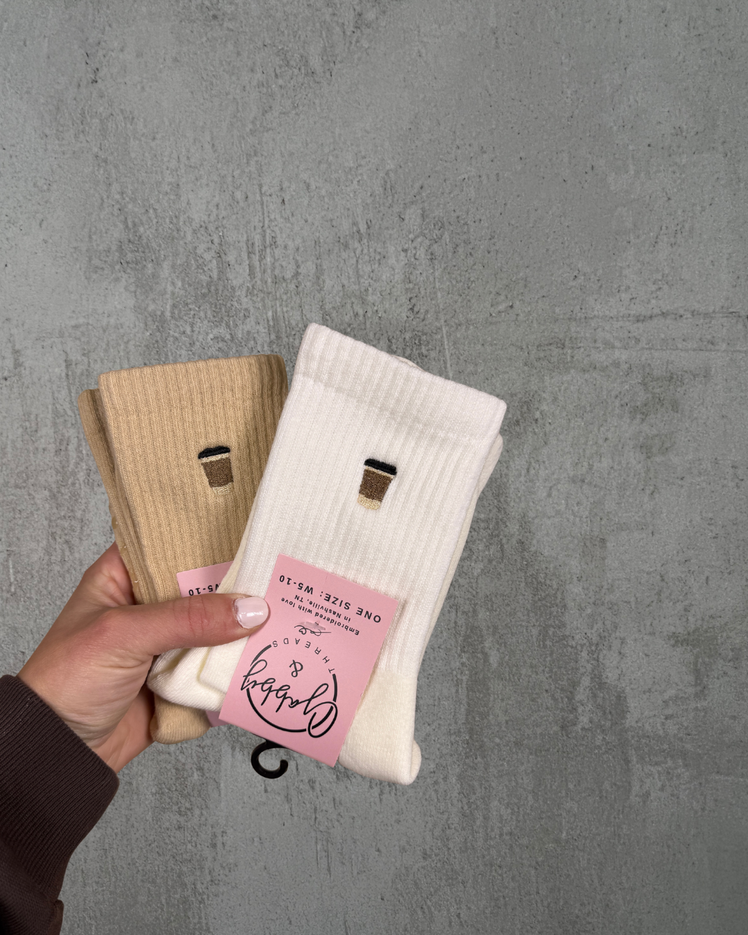 a hand holds up two pairs of socks with an embroidered to go coffee cups on them against a grey background
