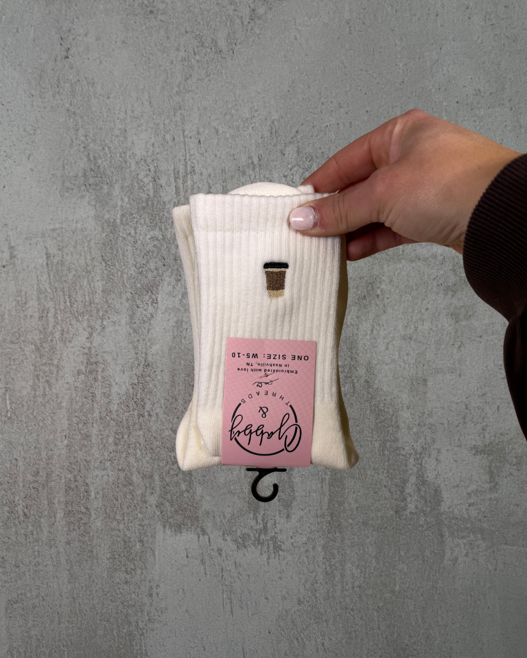 a hand holds up a pair of white grip socks with an embroidered to go coffee cup on it against a grey background