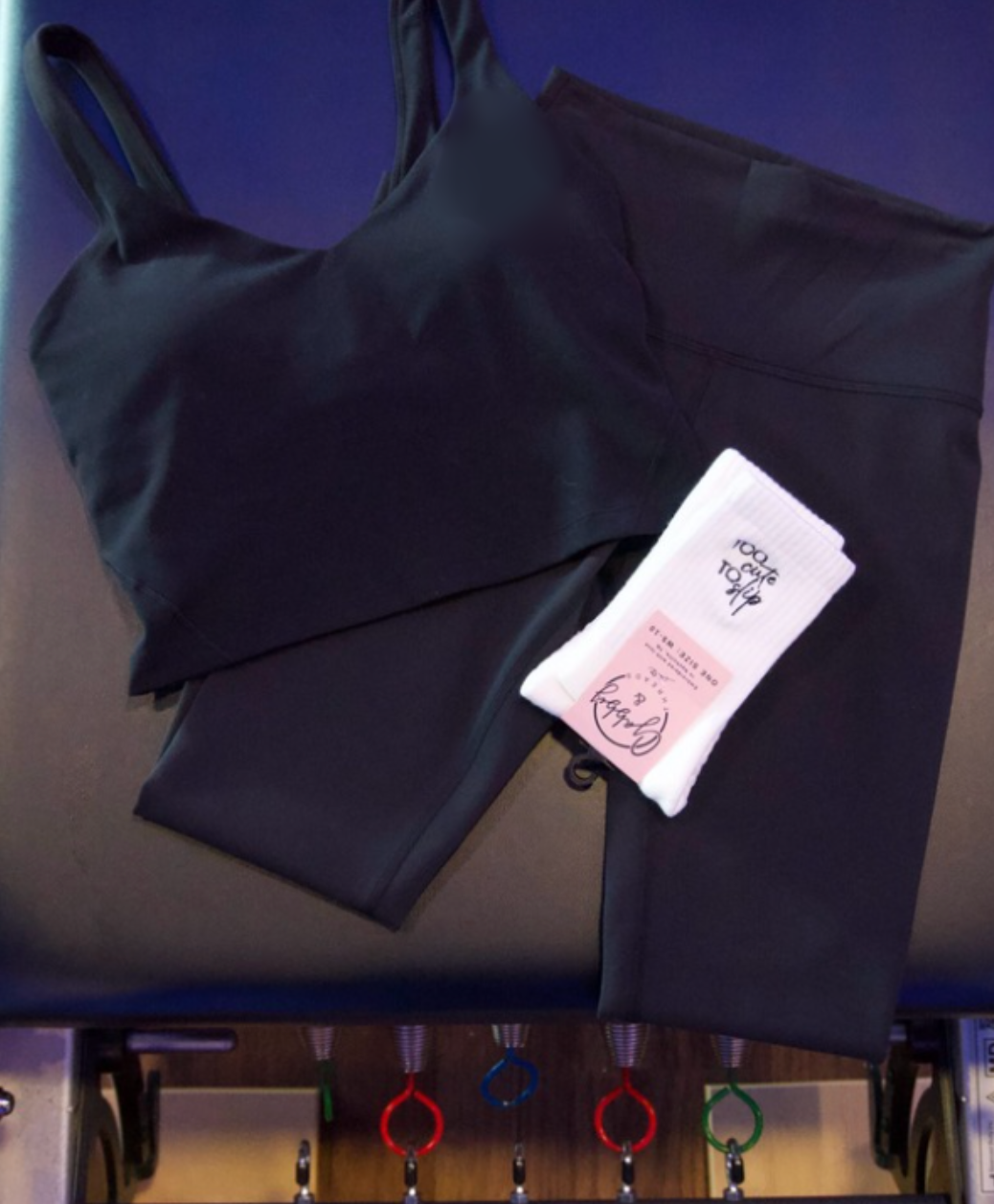a black workout set lays on top of a pilates reformer with a pair of white grip socks that say "too cute to slip"