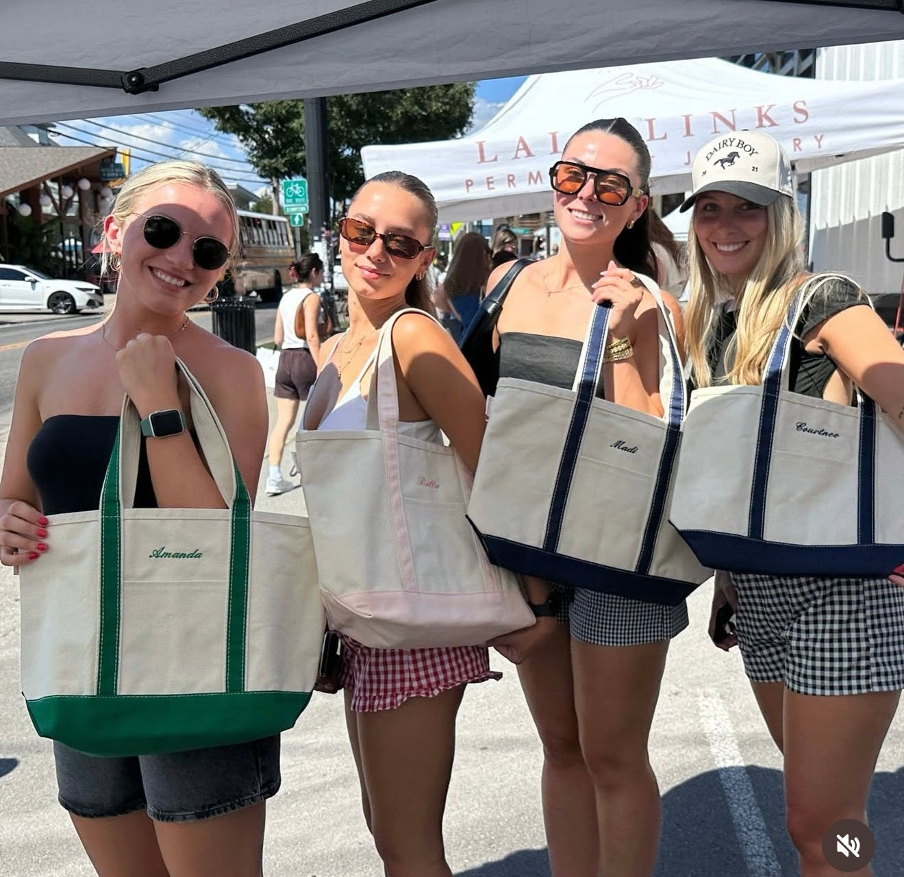 Four women holding branded tote bags outdoors under a tent.