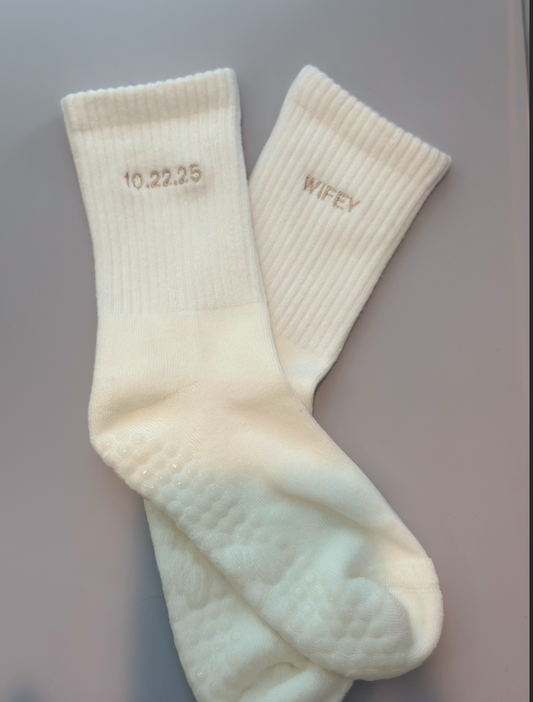 A pair of white grip socks with 'Wifey' embroidered on the outside and the wedding date embroidered on the inside