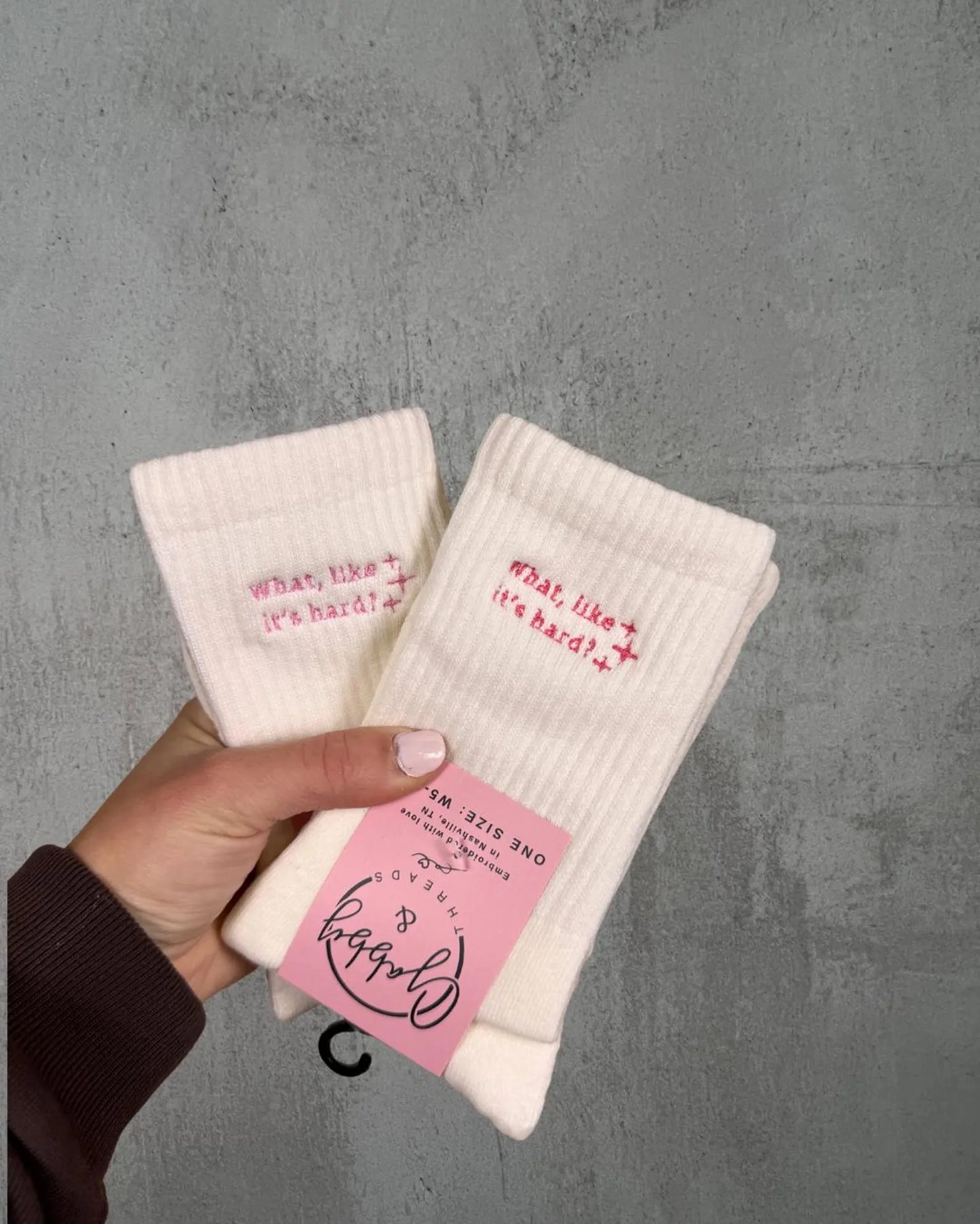 two pairs of white grip socks with the text "What, Like It's Hard" are held up by a hand against a grey background