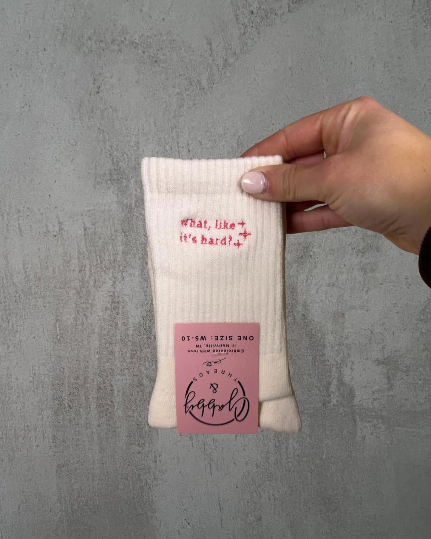 a pair of white grip socks with the text "What, Like It's Hard" are held up by a hand against a grey background