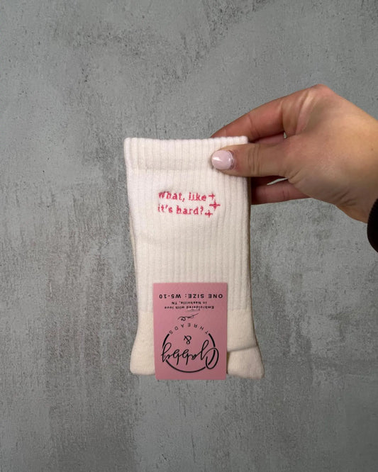 a pair of white grip socks with the text "What, Like It's Hard" are held up by a hand against a grey background