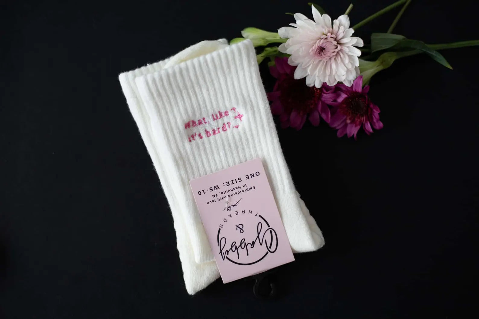a pair of white grip socks with the text "What, Like It's Hard" laying on a black background next to flowers