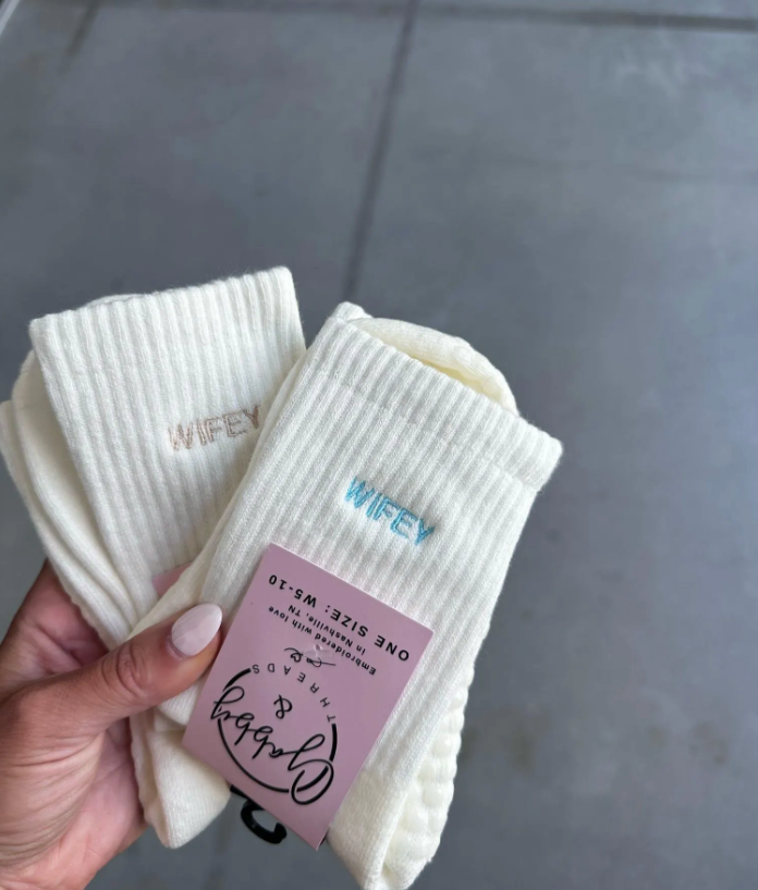 Two pairs of white grip socks with 'WIFEY' text in light blue and beige held by a hand on a gray tiled floor.