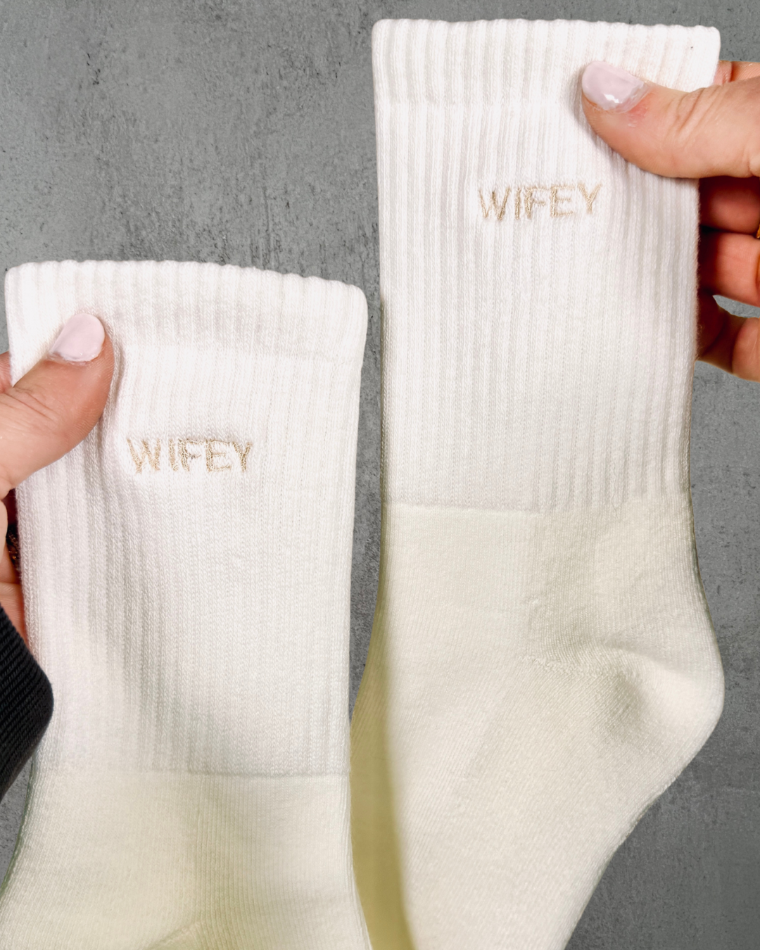 Two white socks with 'WIFEY' embroidery held by a hand against a gray background